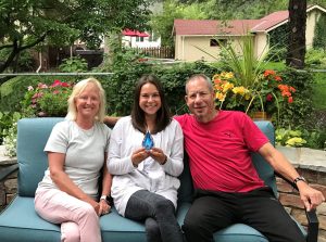 Lori, Sydney, and David sitting on a bench outside. Sydney is sitting between her parents and holding a glass water droplet.